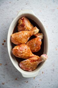 Six seasoned chicken legs in a white casserole dish.