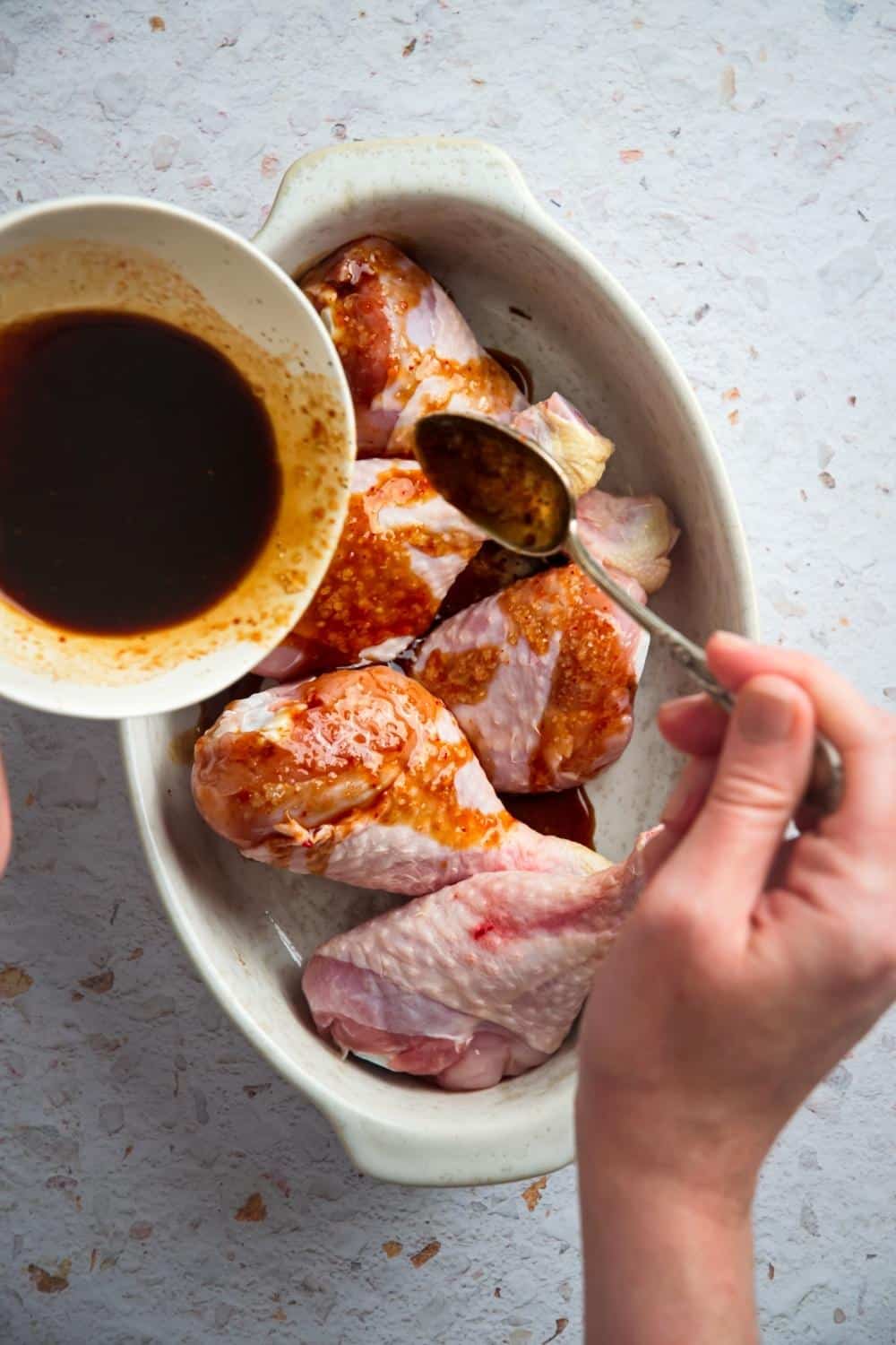 A hand pouring sauce on top of six chicken legs in a casserole dish.