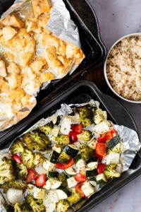 Part of a baking dish with roasted veggies, part of a baking dish with cooked chicken, and part of a bowl of rice.