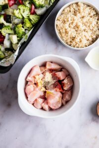 A bowl of cubed raw chicken with seasonings on it. A bowl of rice is behind it.