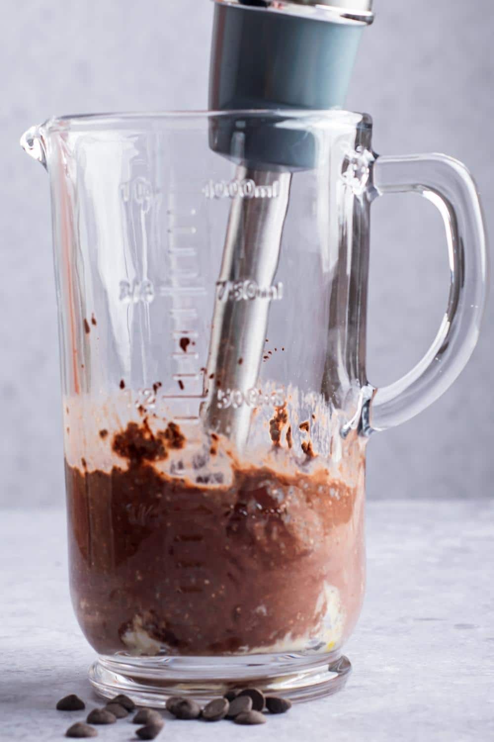 An immersion blender mixing chocolate protein pudding together in a large glass cup.