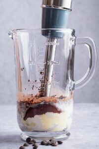 An immersion blender mixing chocolate protein pudding ingredients in a large glass cup.