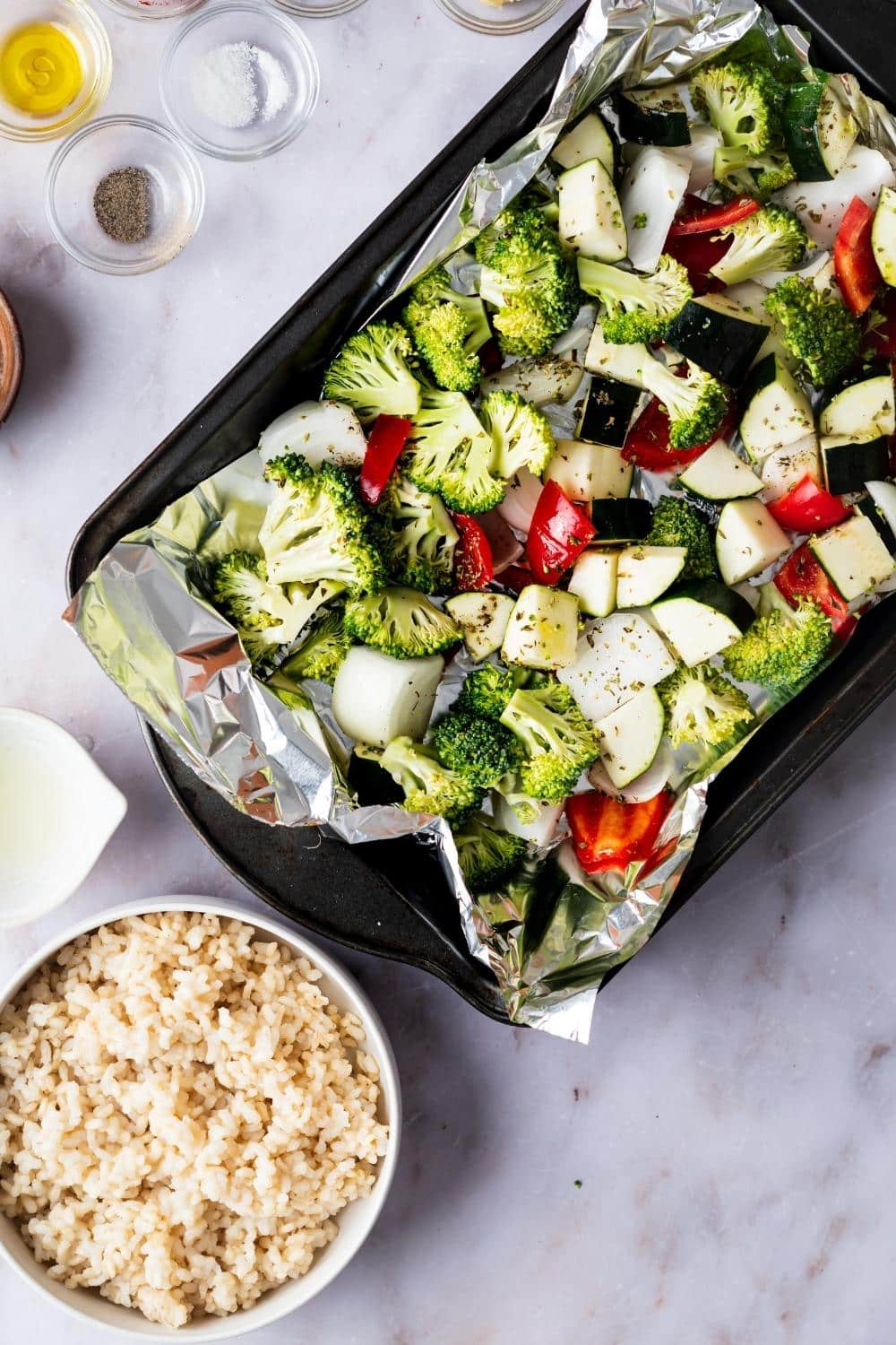 A baking dish with broccoli, zucchini, and bell pepper on it. In front of that is a bowl of rice.