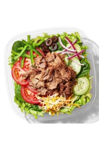 Steak, tomatoes, cucumbers, black olives, and shredded cheese on top of lettuce in a container.