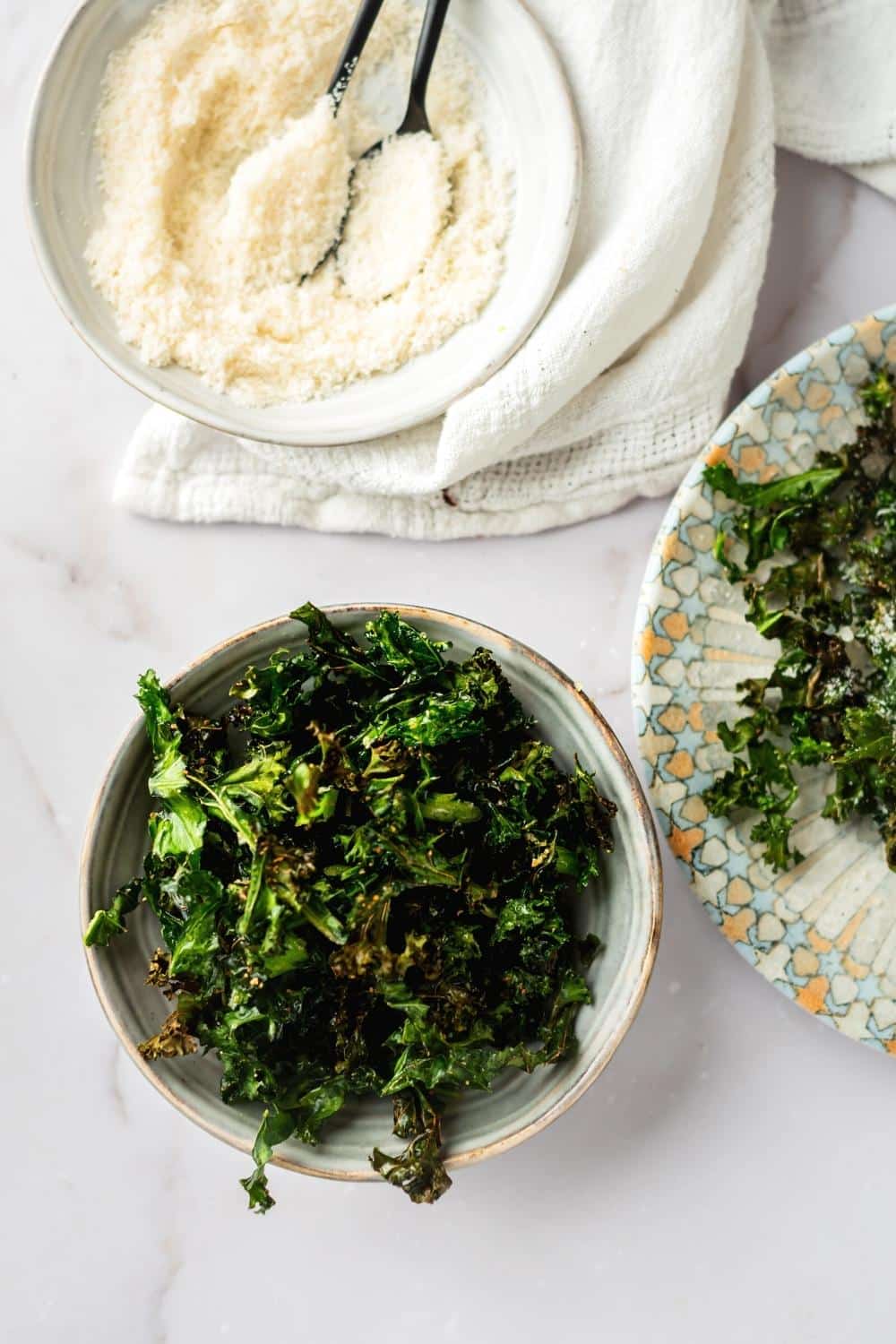 Kale chips in a bowl with a bowl of parmesan cheese behind it.