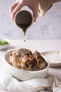 A hand pouring juices on top of a cornish hen in a white casserole dish.