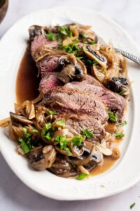 Bottom round roast that is slice with mushrooms and onions surrounding it on a plate.