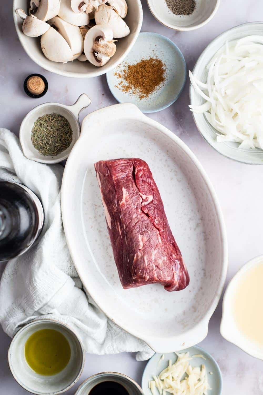 Bottom round roast in a white dish, seasonings in a bowl, cinnamon in a bowl, sliced onions in a bowl, and mushrooms in a bowl on a gray counter.