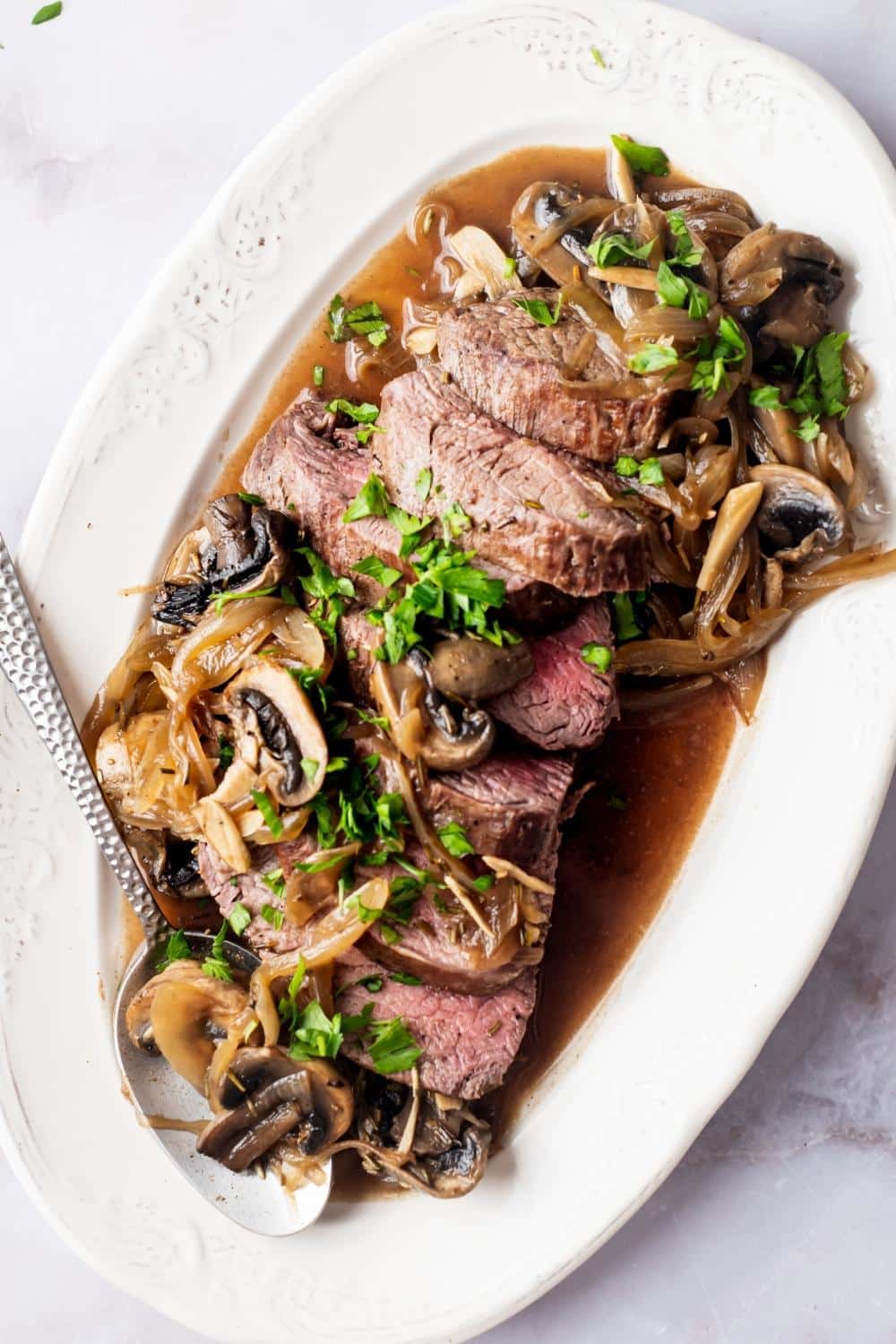 A bottom round roast sliced with mushrooms and onions on top on a white serving plate.