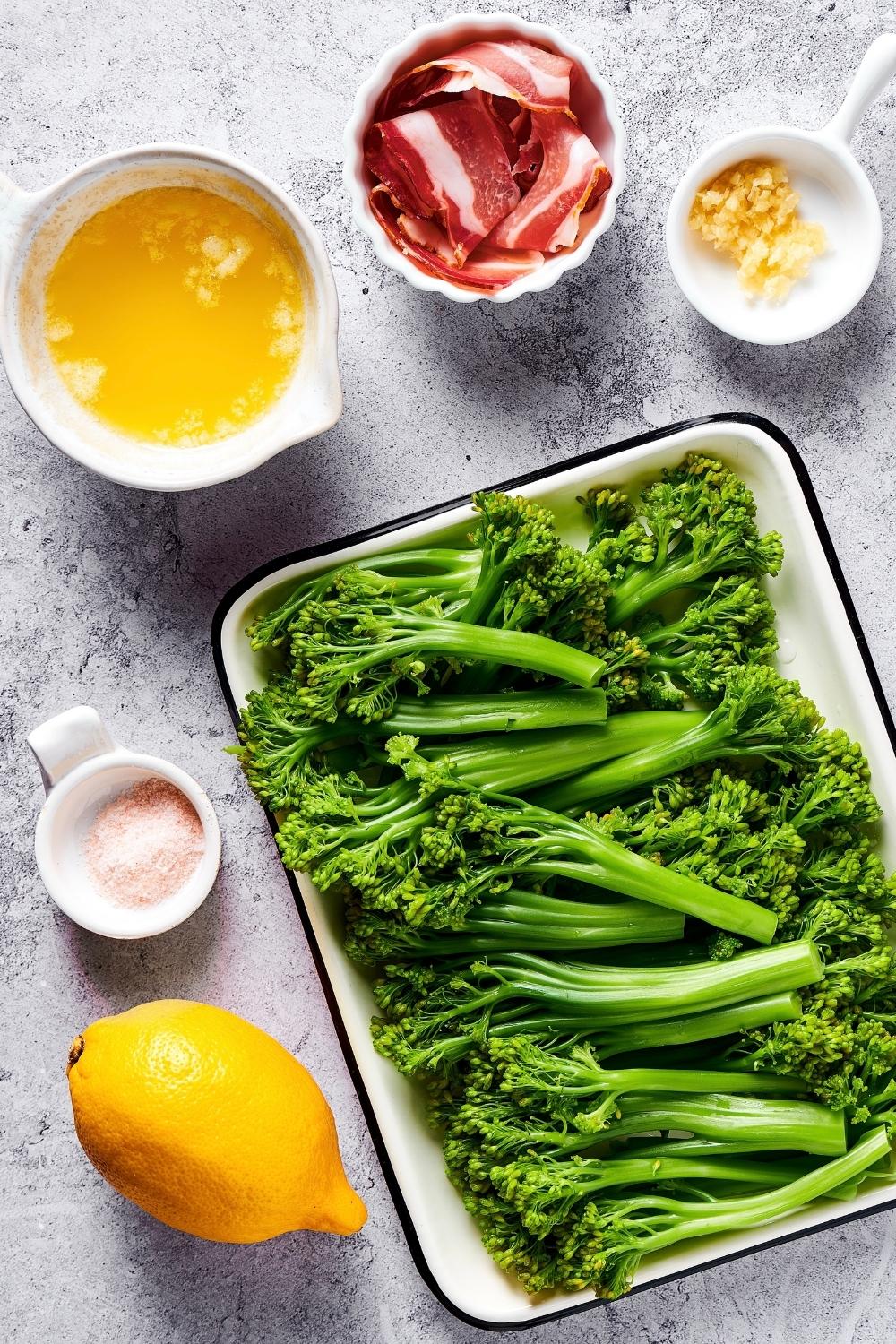 Broccolini on a plate, a lemon, a bowl of salt, a bowl of melted butter, a bowl of bacon, and a bowl of minced garlic.
