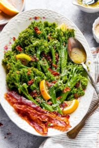 A spoon with lemon garlic butter on it on a plate that has broccolini, lemon wedges, and a few slices of bacon on top.