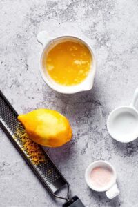 A bowl of lemon juice, a lemon, lemon zest on a grater, a bowl of salt on a grey counter.