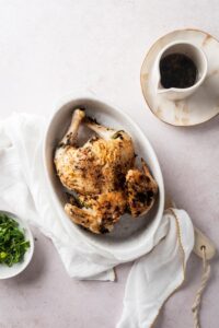 A cornish hen in a casserole dish with a pitcher of cooking juices next to it.