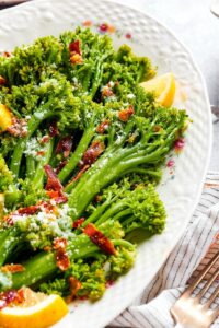 A couple of broccolini stalks with pieces of bacon on part of a white plate.