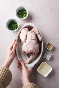 A hand holding a casserole dish with a cornish hen in it. Surrounding it it a bowl of oil, a bowl of cilantro, and a bowl of parsley.