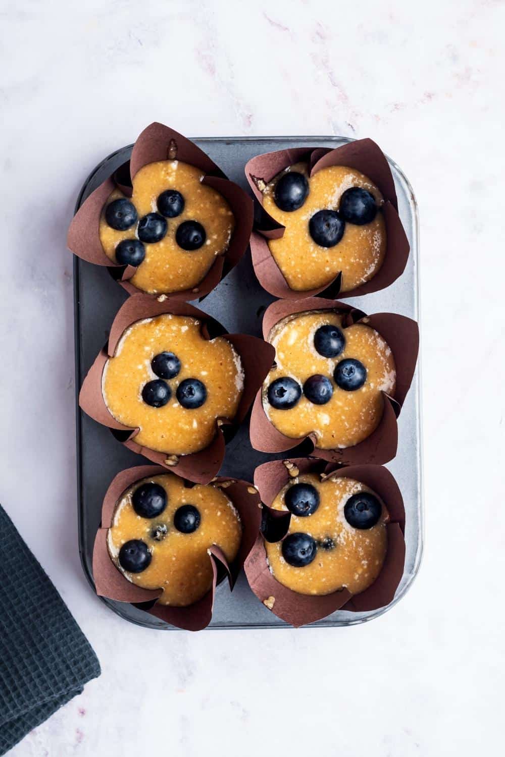 Blueberry muffin batter and six muffin cups in a muffin tin.