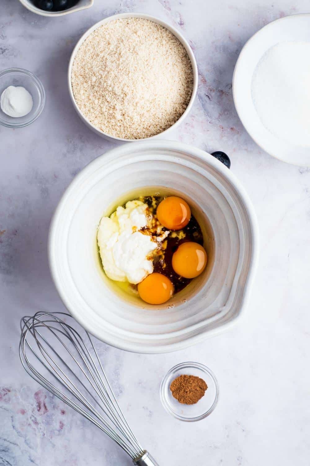 Three eggs, sour cream, and vanilla extract in a white ball. Behind that is a bowl of almond flour.