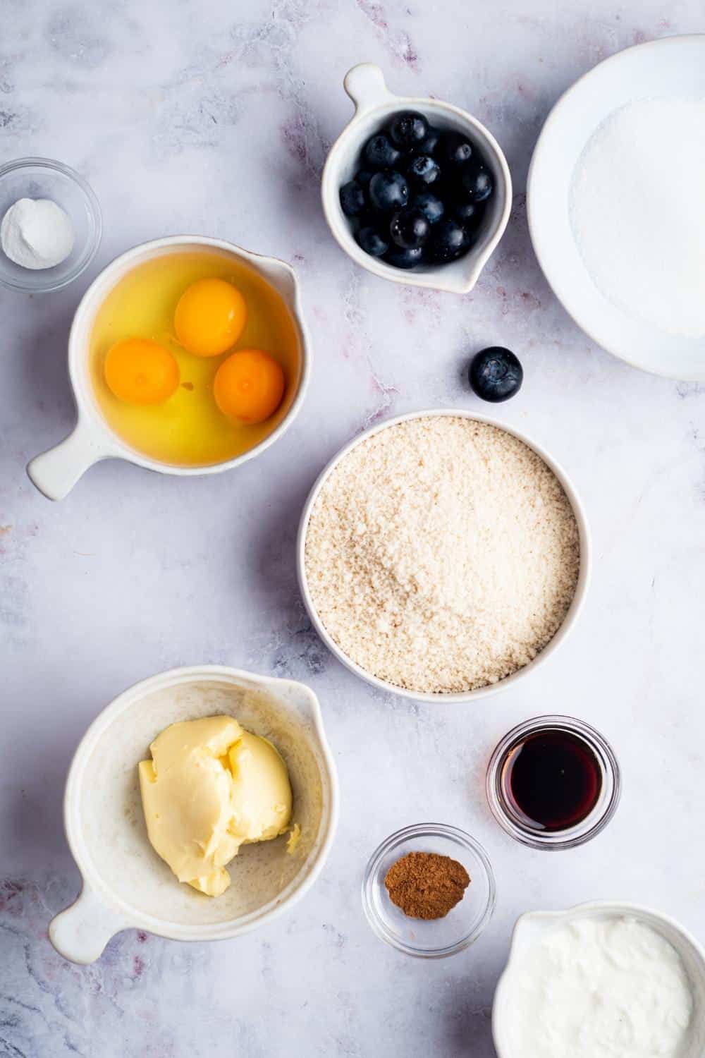 A bowl of three eggs, a bowl of blueberries, a bowl of flour, a bowl of sour cream, and a bowl of powdered sugar all on a gray counter.