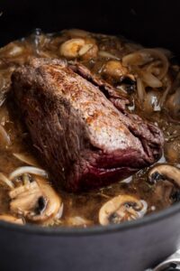 Bottom round roast cooking in a Dutch oven with mushrooms and onions in it.