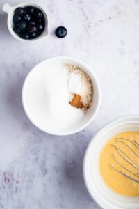 A bowl of dry ingredients for blueberry muffins, part of a bowl of the wet ingredients, and above blueberries on a gray counter