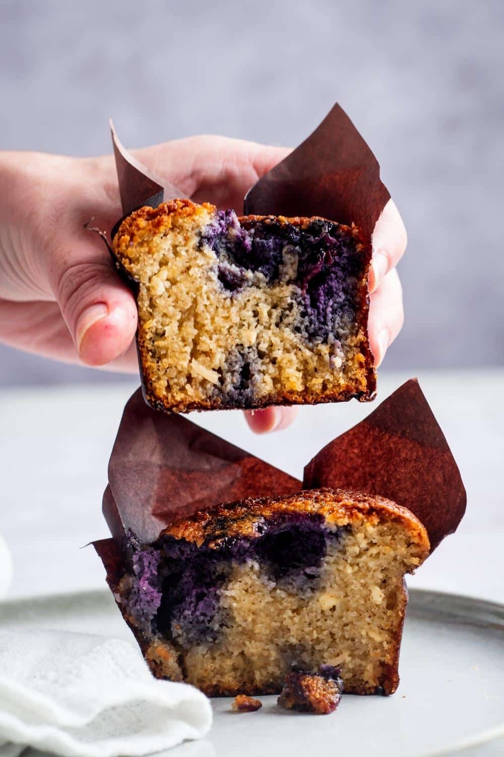 A hand holding half of a blueberry muffin over another half of the muffin.