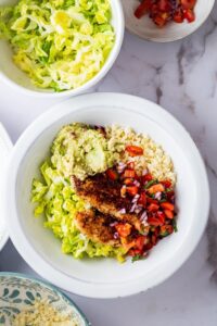 Chicken, lettuce, guac, pico, and cauliflower rice in a bowl. Behind that is a bowl of lettuce.