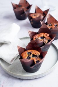 Six blueberry muffins in muffin tins in a row on the counter and plate.