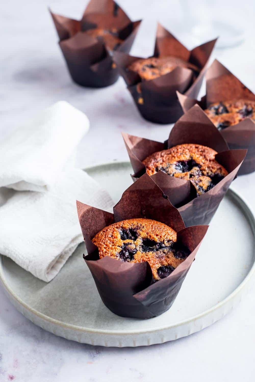 Six blueberry muffins in muffin tins in a row on the counter and plate.