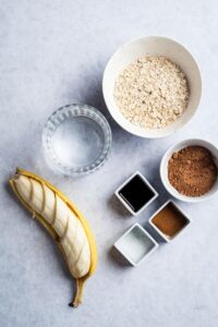 A sliced banana, a bowl of oats, a bowl of protein powder, a square bowl of vanilla extract, and a bowl of water on a grey counter.