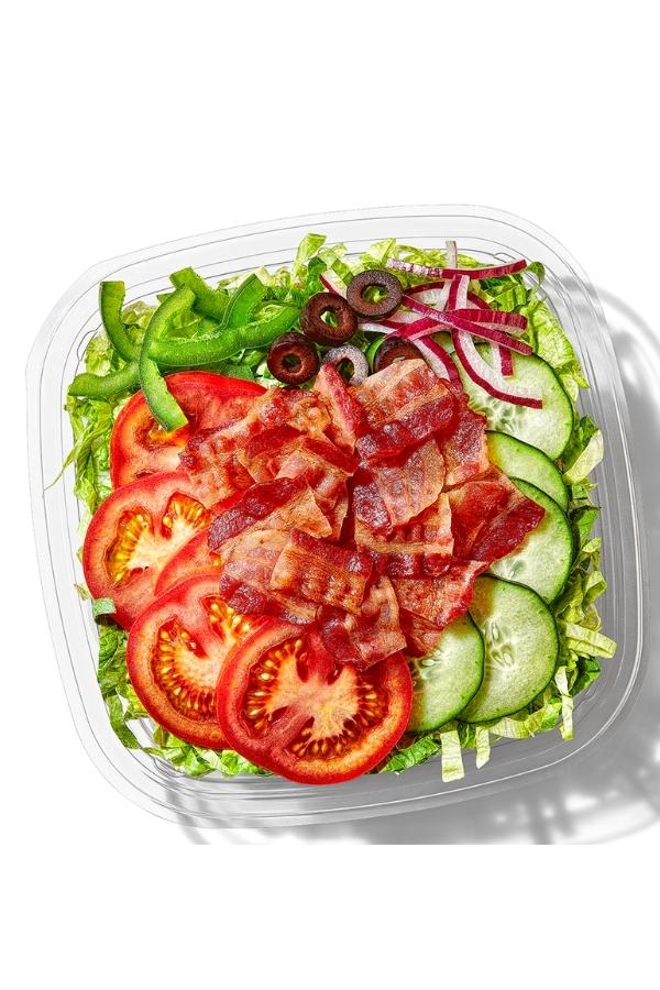 Bacon, tomato, cucumbers, red and green onion, and black olives on top of lettuce in a conatiner.