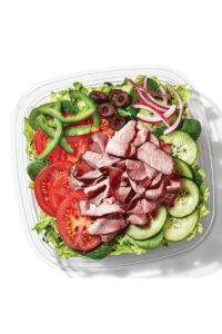 Roast beef, cucumbers, tomatoes, olives, green peppers, and red onion on top of lettuce in a container.