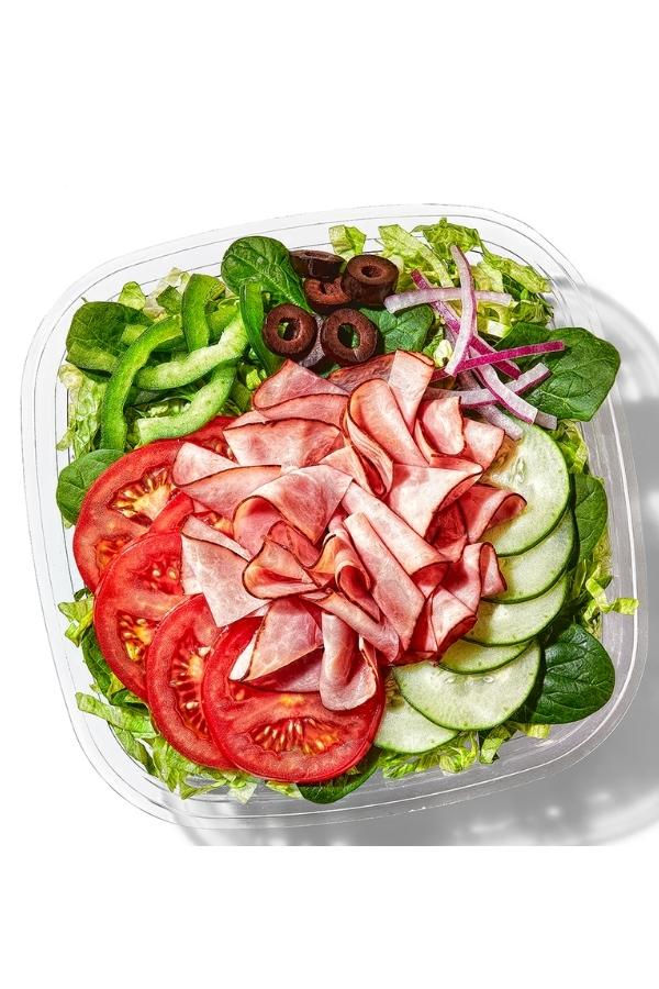 Ham, cucumbers, tomatoes, black olives, red onion, green onion on top of lettuce in a container.