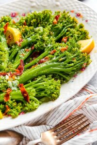 A bunch of broccolini on part of a white plate with some chopped bacon on it.