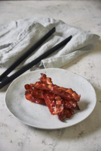 A stack of baked turkey bacon on a white speckled plate. A tea towel and a pair of black tongs are in the background.