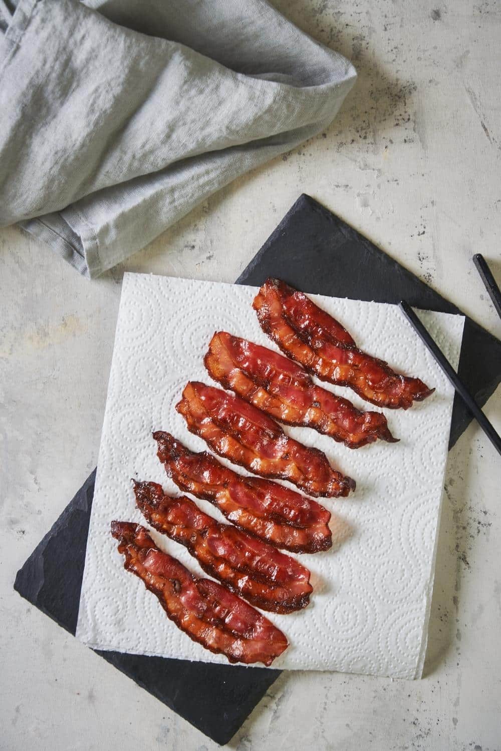 Six pieces of cooked turkey bacon on a paper towel on top of a slate serving platter. A pair of black tong rests on the edge of the platter and a tea towel is folded on the upper left corner.