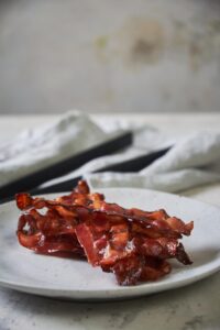 A stack of cooked turkey bacon on a speckled white plate. A pair of black tongs on top of a folded tea towel can be seen in the background.
