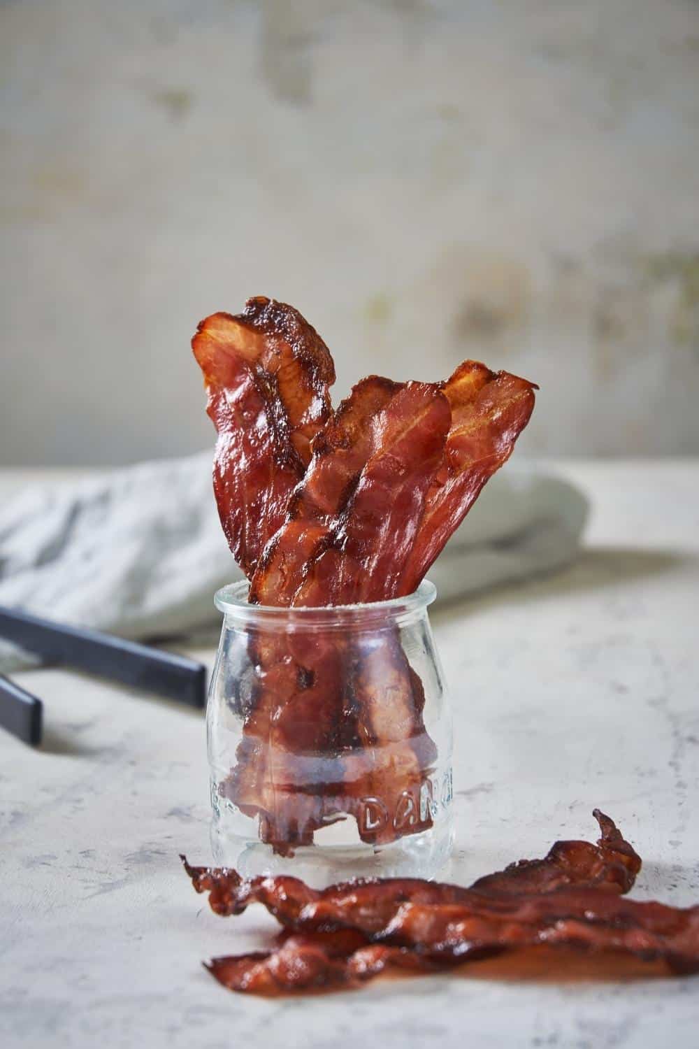 Cooked turkey bacon slices standing upright in a small glass jar. Two more slices of bacon sit outside the jar on the marble countertop. Black tongs and a tea towel are in the back.