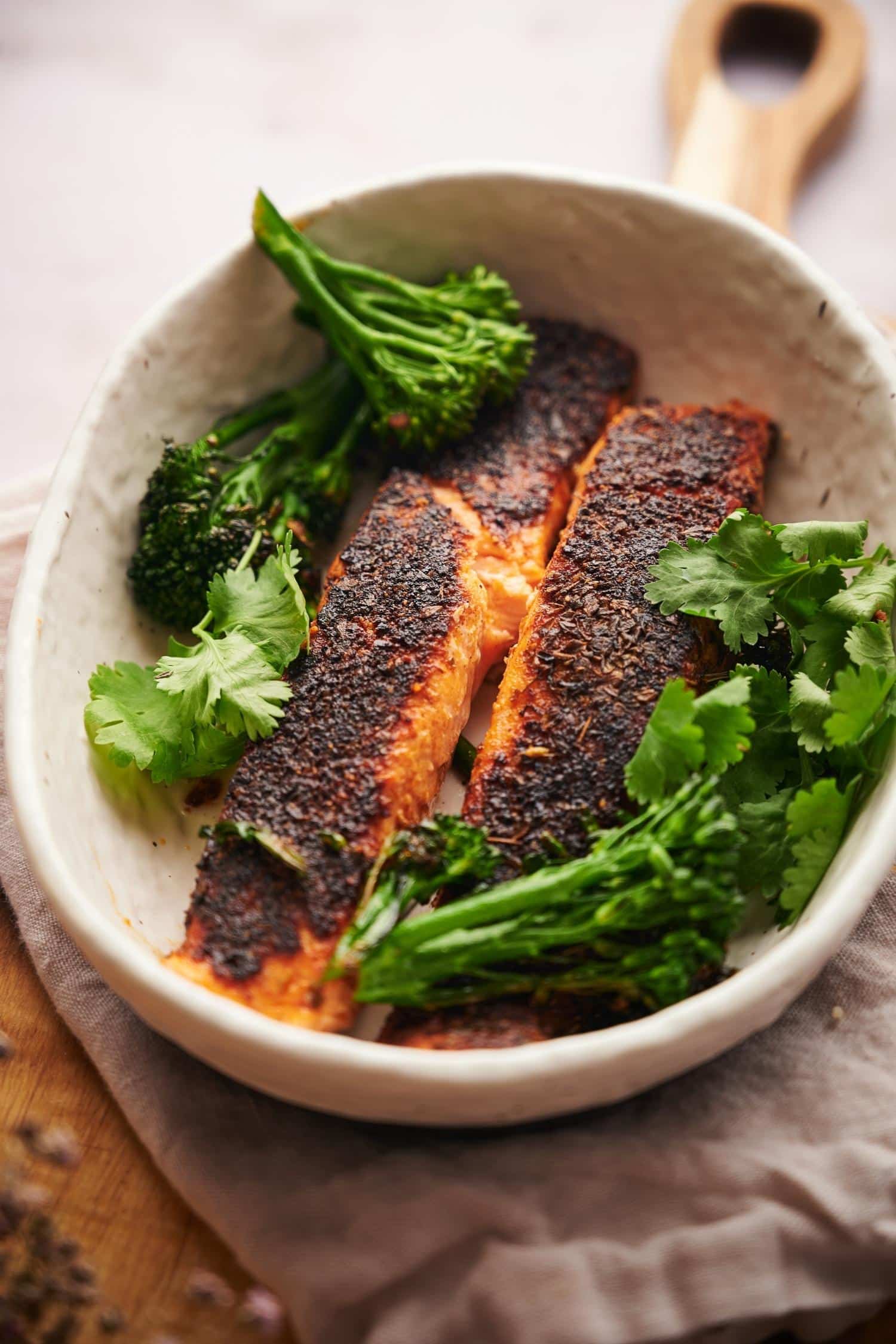 two blackened salmon fillets in a shallow white bowl garnished with seared broccolini and fresh cilantro.