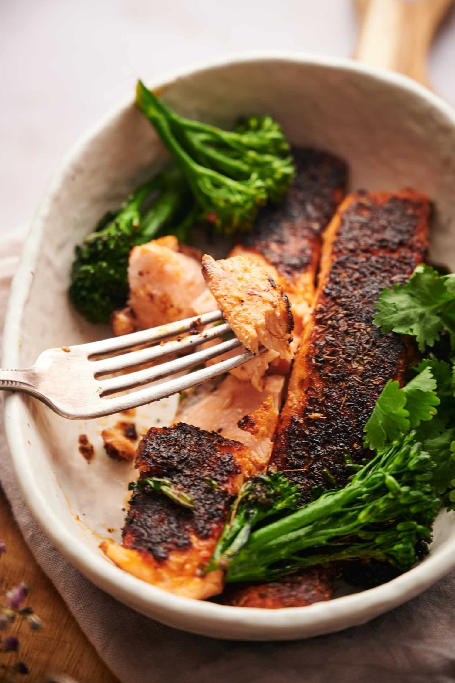 A fork taking a piece of blackened salmon. The salmon is pink and flakey, served in a shallow white bowl with seared broccolini and fresh cilantro for garnish.