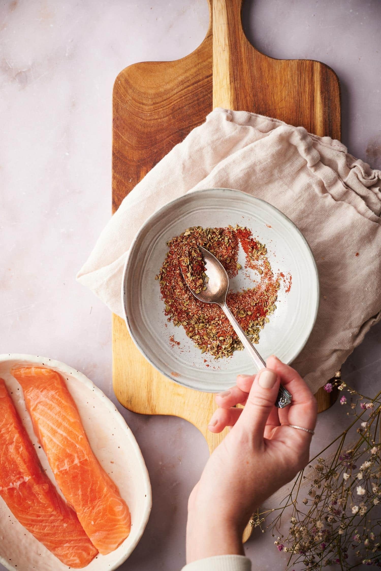 A hand stirring the blackening seasoning mix together in a bowl with a metal spoon. To the side is two raw salmon fillets.