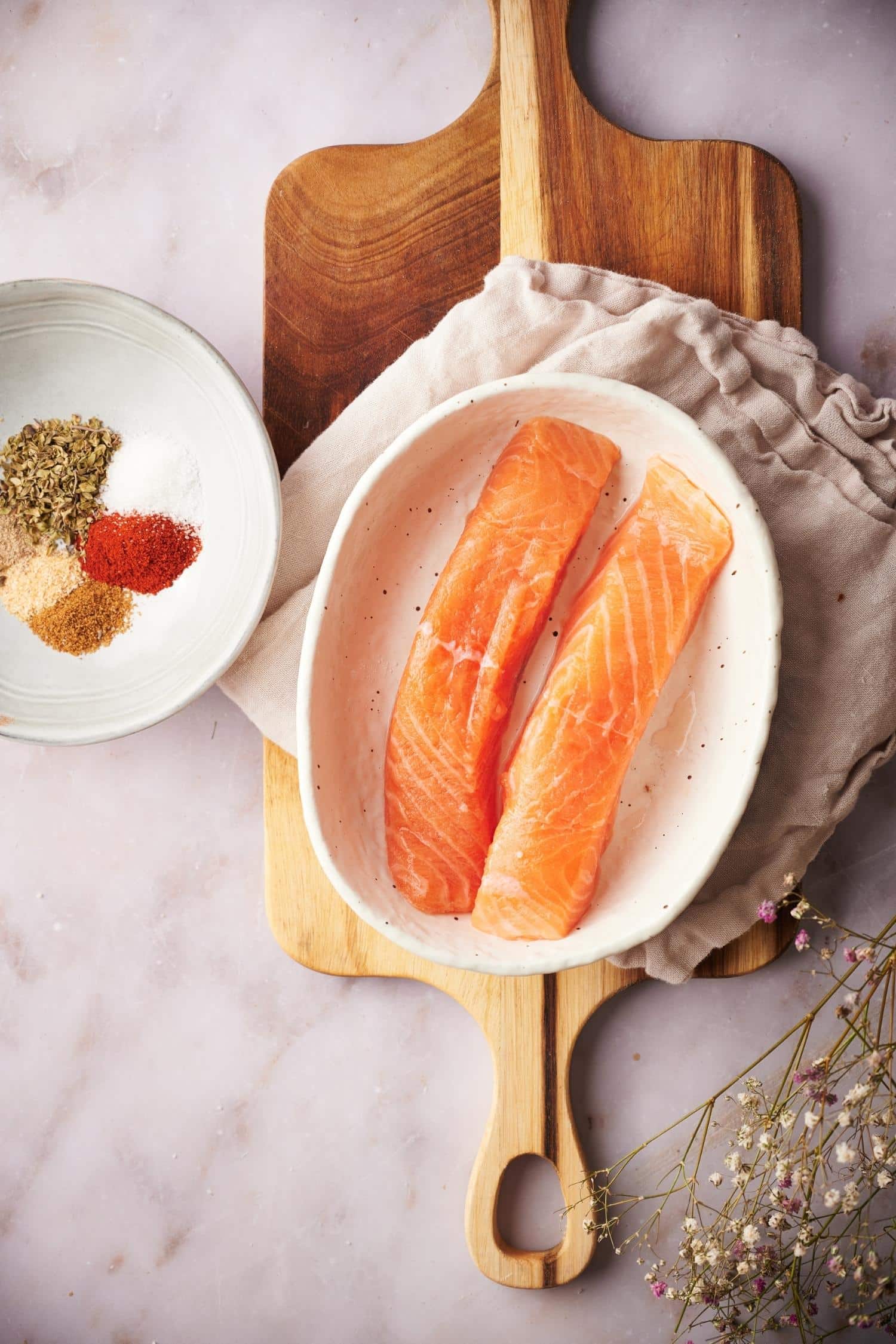 Two raw salmon fillets in a shallow bowl resting on top of a tea towel on a wooden cutting board. To the side is a bowl of spices that haven't been combined yet.