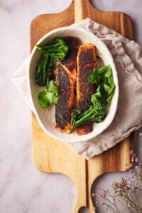 Top shot of two salmon fillets with seared broccolini and fresh cilantro in a shallow oval bowl. The bowl is resting on top of a tea towel laid on a wooden cutting board.