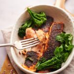 Two blackened salmon fillets in a bowl with seared broccolini and fresh cilantro. A metal fork is taking a piece of flakey salmon.