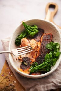 Two blackened salmon fillets in a bowl with seared broccolini and fresh cilantro. A metal fork is taking a piece of flakey salmon.
