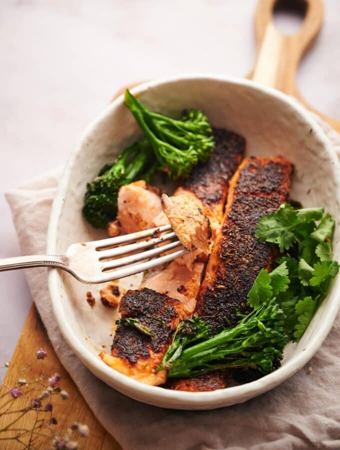 Two blackened salmon fillets in a bowl with seared broccolini and fresh cilantro. A metal fork is taking a piece of flakey salmon.