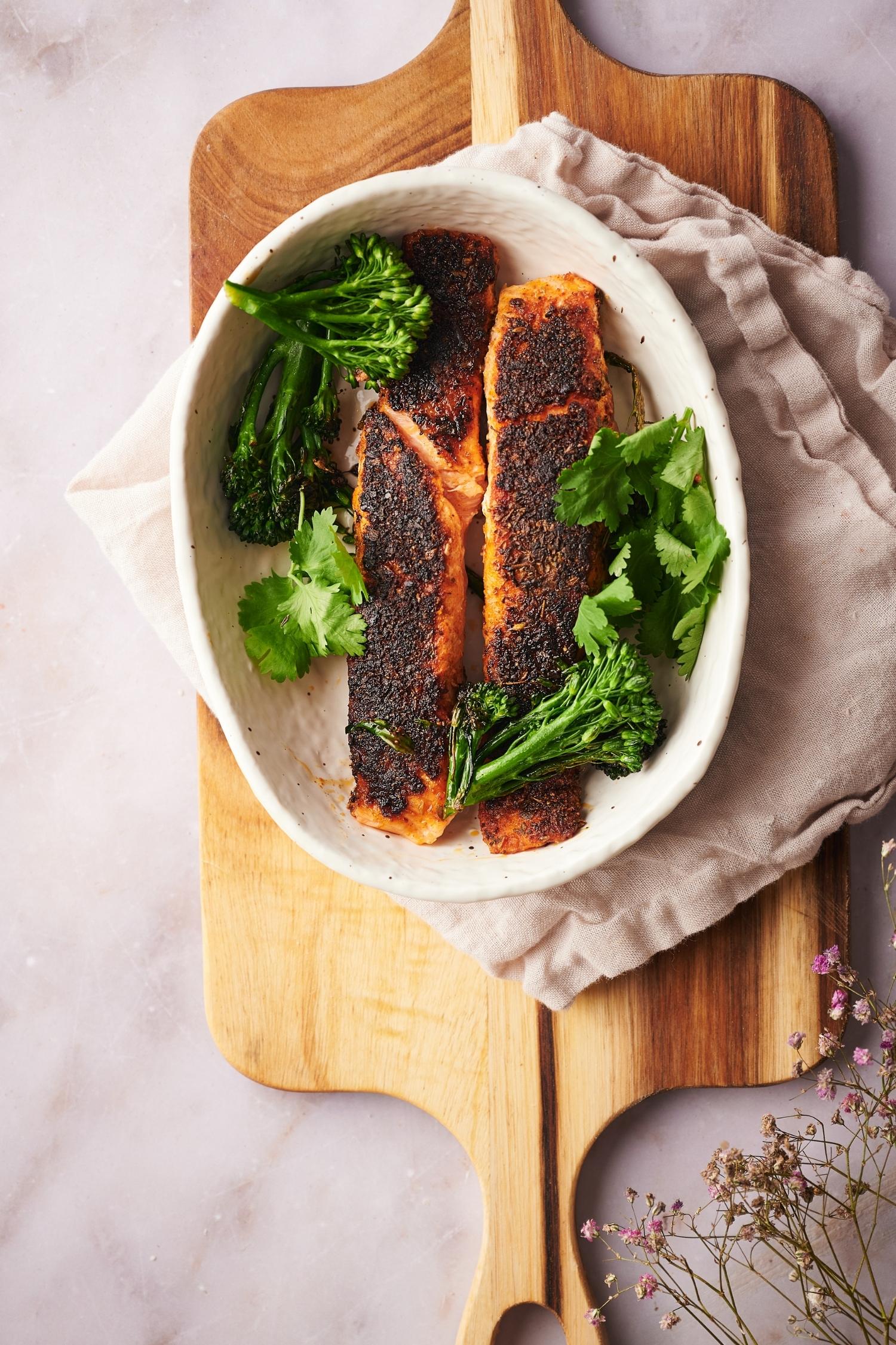 Top shot of two salmon fillets with seared broccolini and fresh cilantro in a shallow oval bowl. The bowl is resting on top of a tea towel laid on a wooden cutting board.