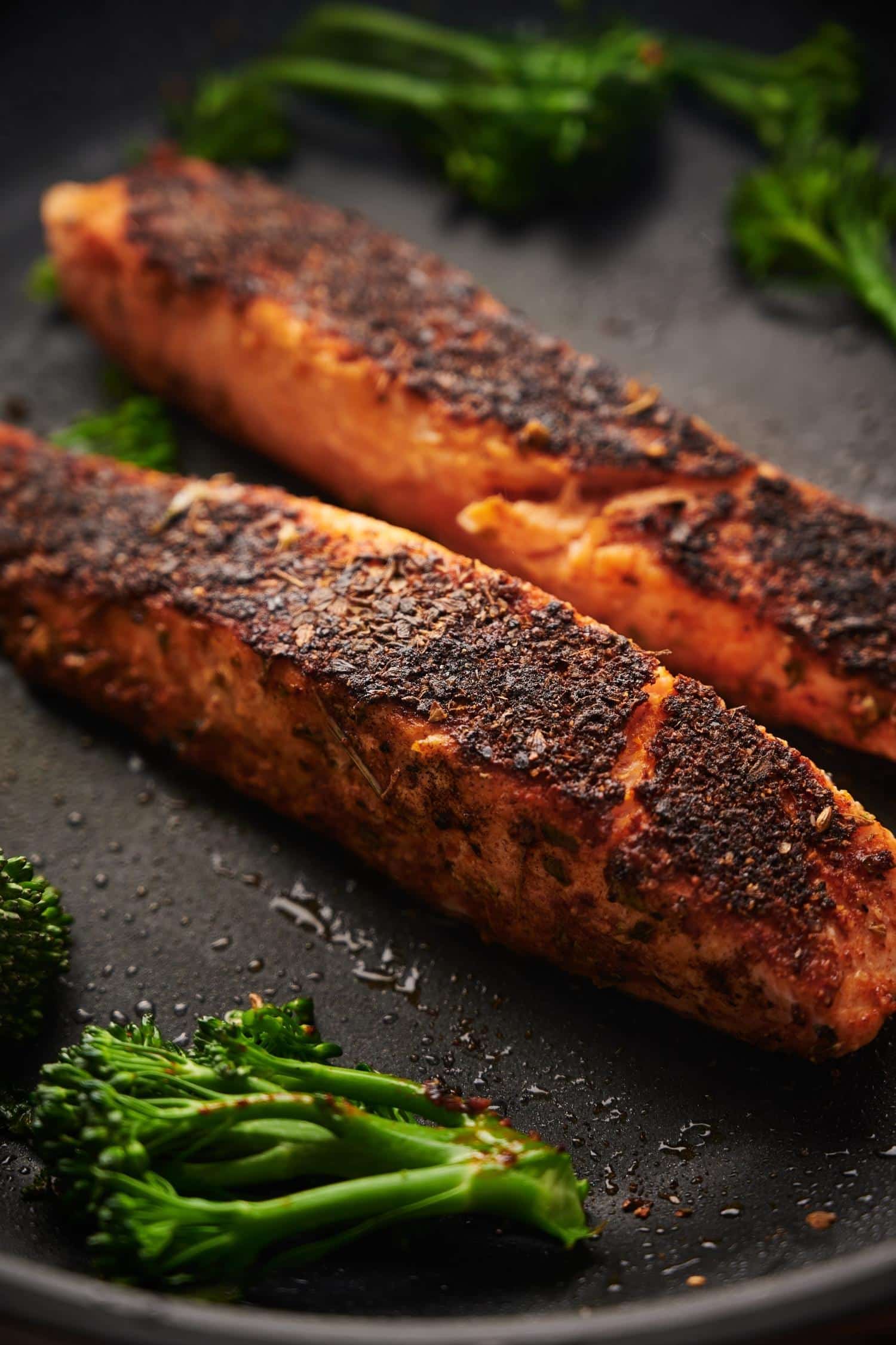 Close up of blackened salmon fillets in a pan. The topside of the salmon is crisp and blackened and they're surrounded by pieces of broccolini.