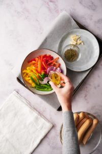 Olive oil being drizzled on a bowl of sliced bell peppers and onions. To the side is a plate of sliced garlic with a small bowl of herbs.