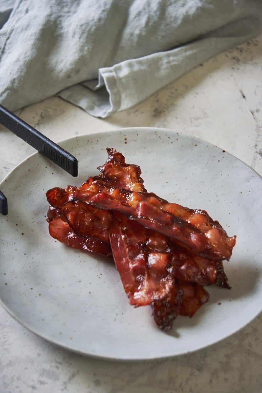 A plate of 5 cooked turkey bacon slices stacked on top of each other on a speckled white plate. A pair of black tongs sit on the edge of the plate and a white tea towel rests in the back.