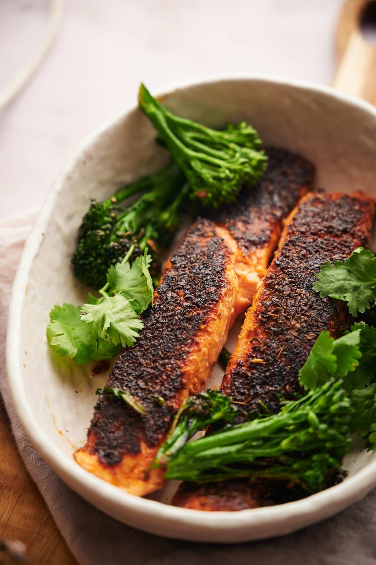 Two blackened salmon fillets in a shallow white bowl with seared broccolini and fresh cilantro for garnish.